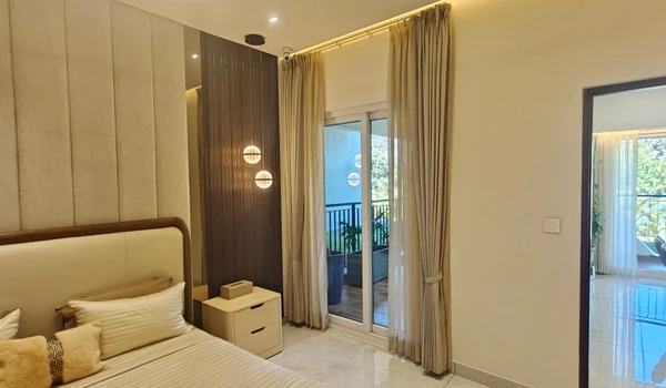 Luxurious master bedroom at Nambiar District 25 4 BHK model flat, featuring a beige upholstered headboard, pendant lighting, and large sliding glass doors opening to a private balcony
