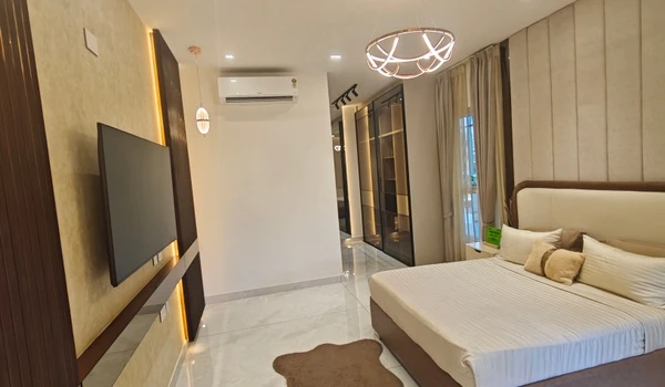 Spacious master bedroom in the Nambiar District 25 4 BHK model flat, showcasing a modern TV unit with backlighting, a designer chandelier, and a walk-in glass wardrobe