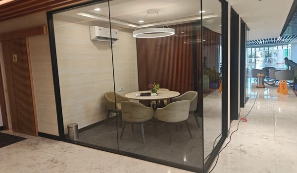 Glass-enclosed private meeting room at Nambiar District 25 marketing office featuring a round discussion table, circular pendant light, and wooden slat wall accents