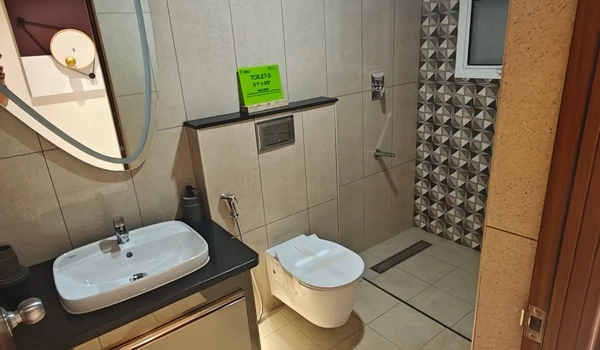 Modern bathroom interior at Nambiar District 25 model flat featuring a wall-mounted commode, sleek vanity washbasin with black countertop, backlit oval mirror, and geometric shower wall tiles