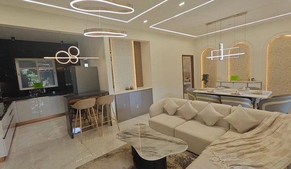 Panoramic interior view of the Nambiar District 25 model flat, showcasing a luxury integrated living space with a white sectional sofa, designer marble coffee table, modern breakfast bar, and open kitchen with high-end light fixtures
